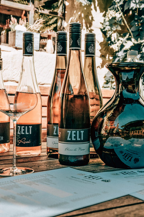 Einige frisch gekühlte Flaschen Rosé und Blanc de Noir Flaschen stehen in der Sonne auf einem Holztisch. Davor steht ein Weinglas. Im Hintergrund sind Pflanzen und das Gebäude des Weingut Zelt zu sehen.